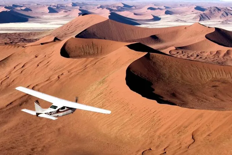 A small aircraft flies above the red dunes of the Namib Desert.