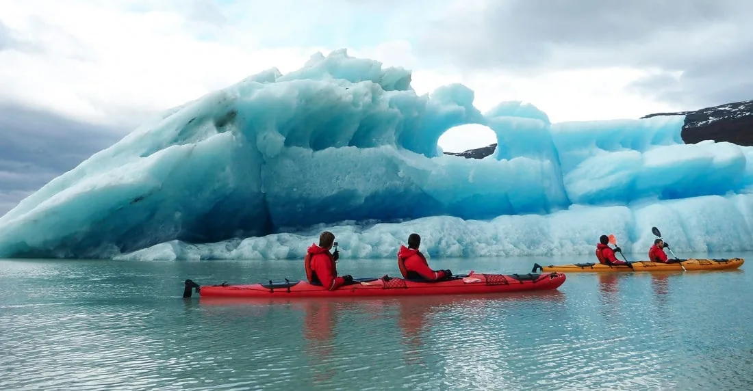 Kayakers paddle across a turquoise lake between floating icebergs.