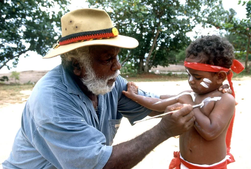 An adult in a broad-brimmed hat gently paints a young person's face outdoors.
