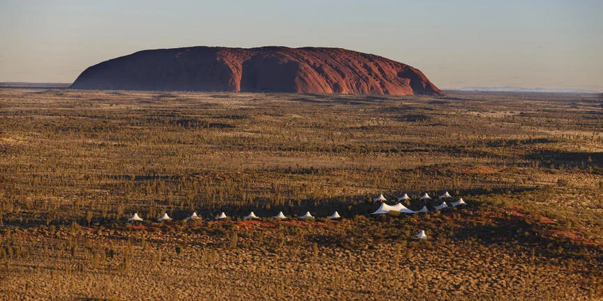 Uluru rising above red desert with a line of white tents in the foreground.