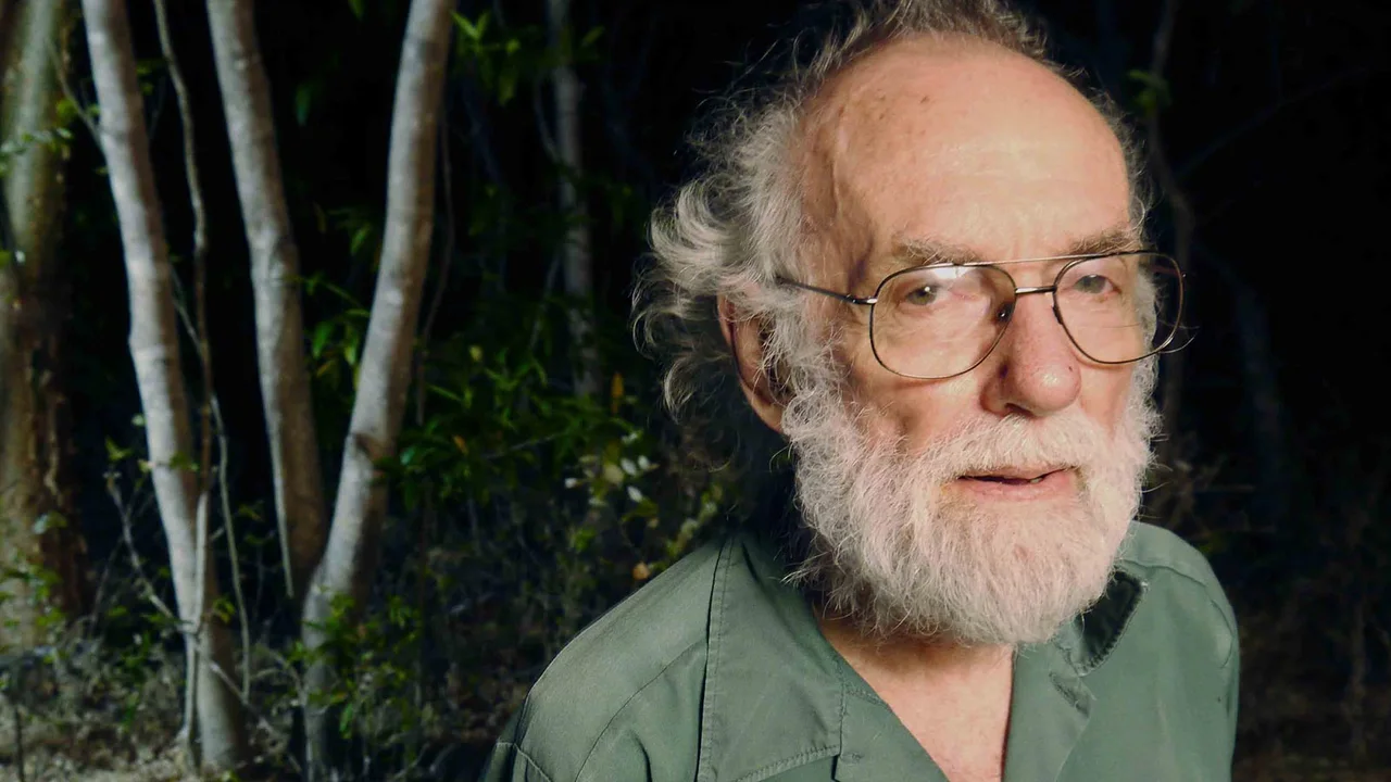 A white-bearded man wearing glasses and a green shirt in a forest.