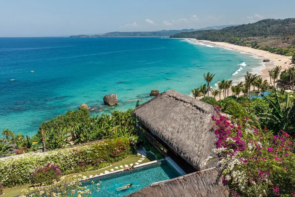 A thatched villa and pool overlooking a palm-lined beach and turquoise sea.