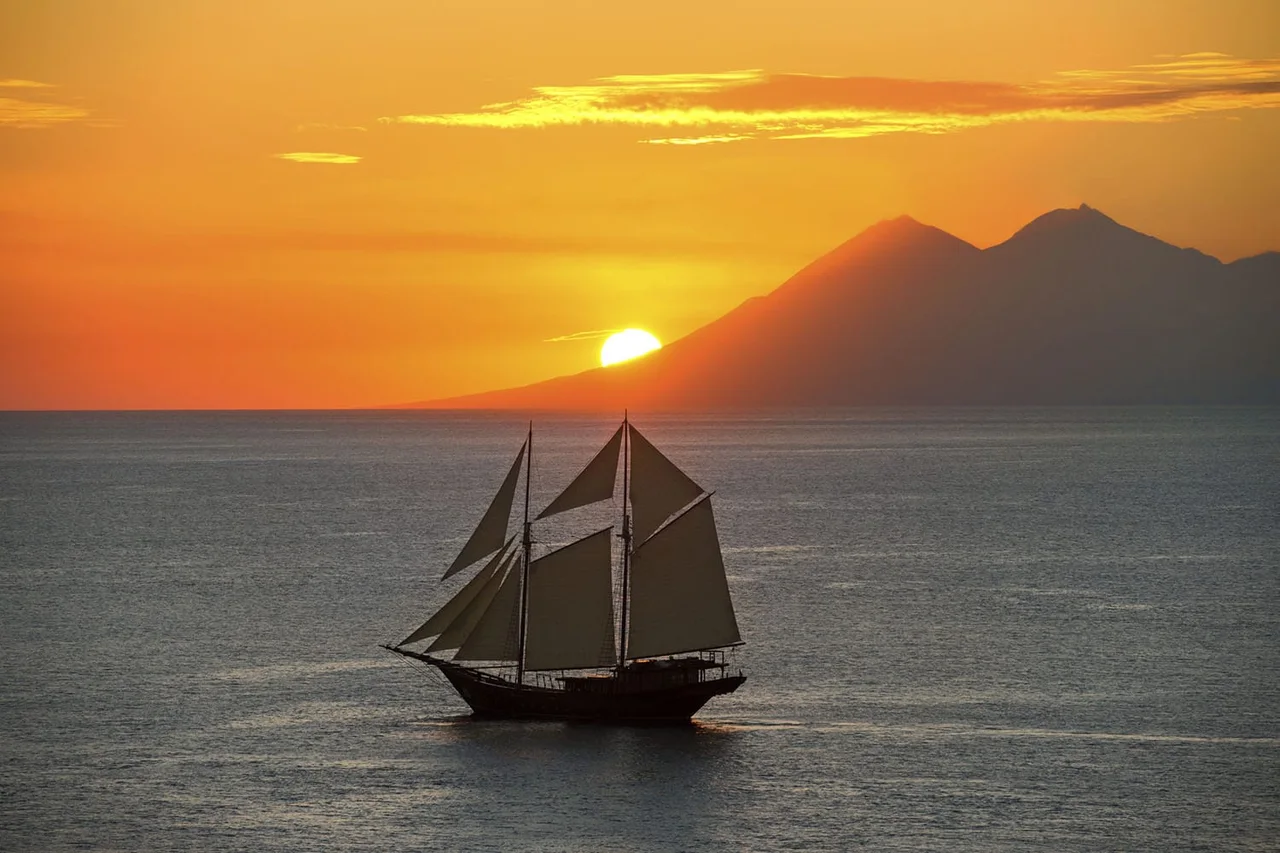 A traditional sailing boat on calm water beneath an orange sunset and mountainous coastline.
