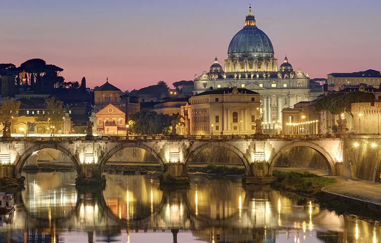 St Peter's dome and illuminated riverside buildings reflected in the Tiber at dusk.