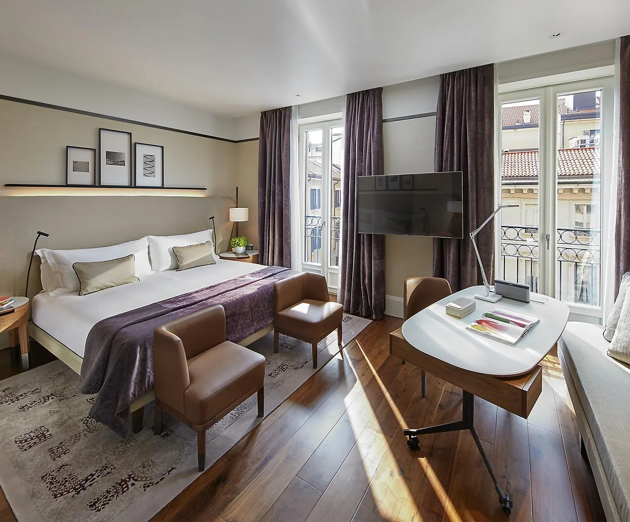 A hotel bedroom with a sofa, desk and tall windows overlooking city buildings.