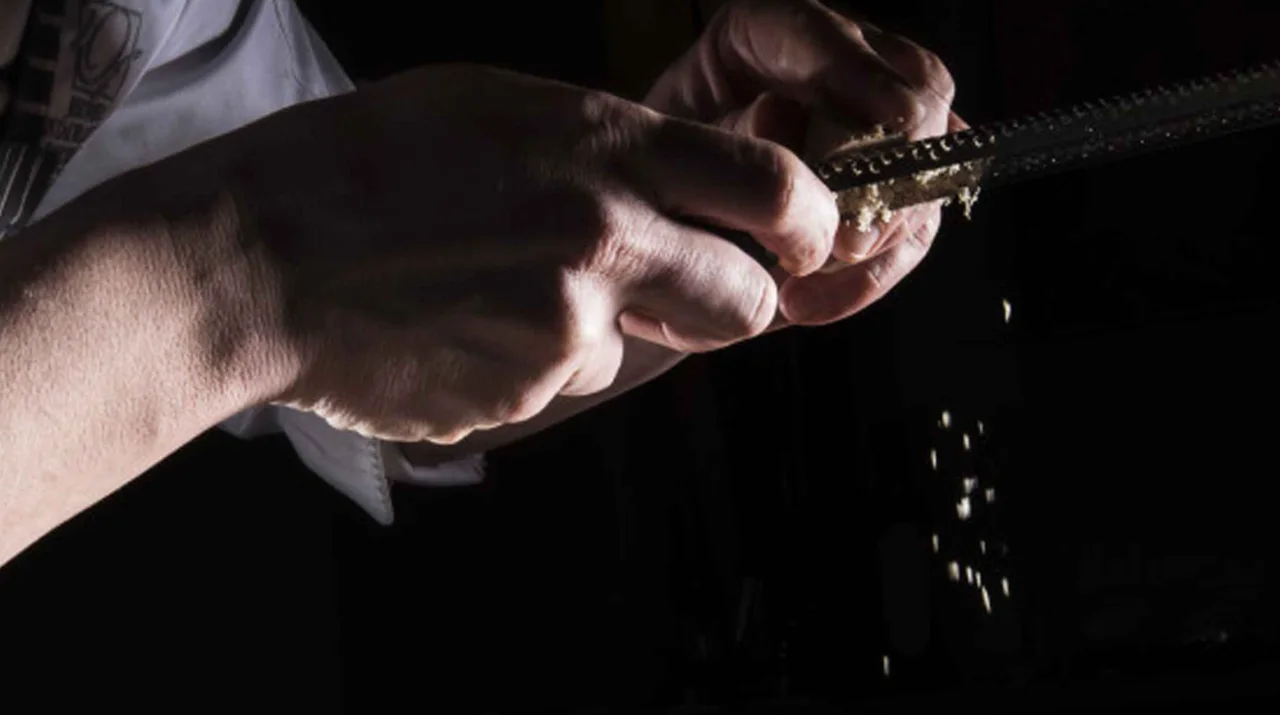 Close-up of hands using a fine grater, with shavings falling against a dark background.