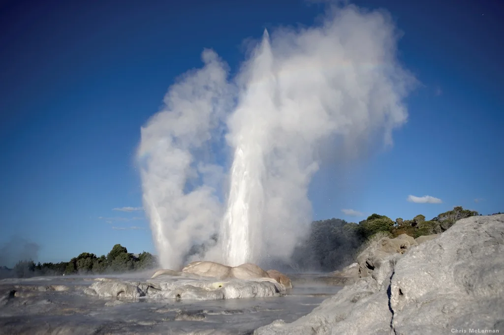 A geyser sending a plume of steam above rocky geothermal terrain.