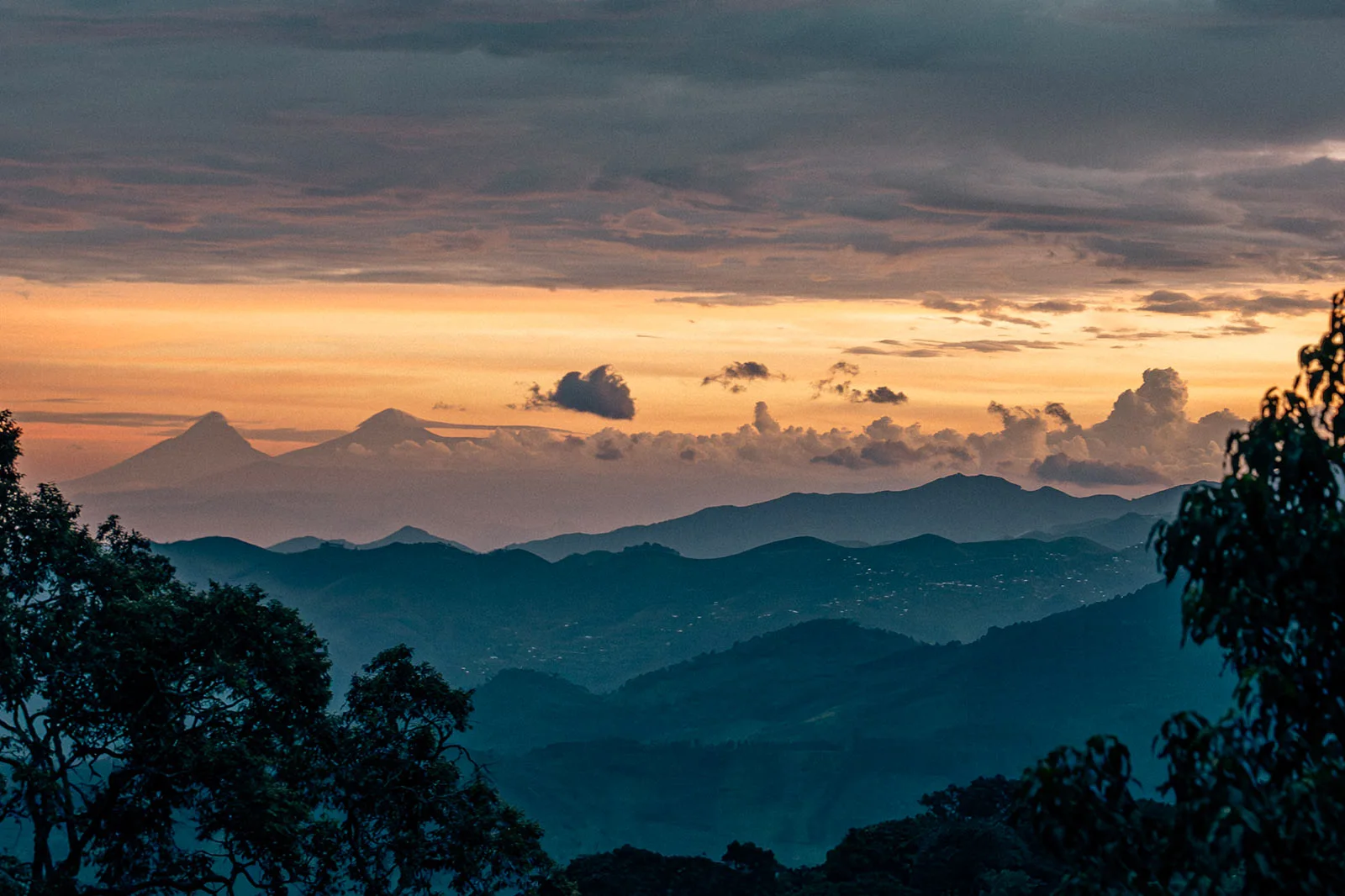 Layered mountain ridges and forested hills beneath an orange sunset.