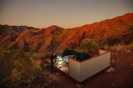 An open-air sleeping shelter sits among the red rocky Flinders Ranges at dusk.