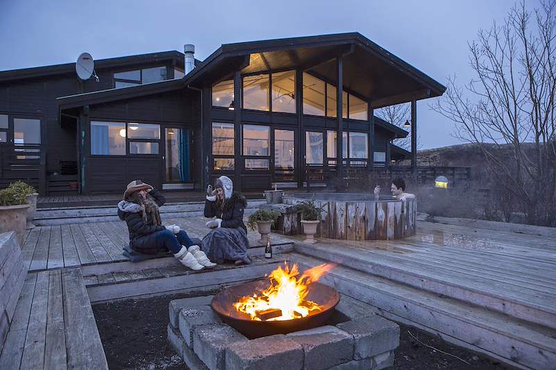 Guests gather around an outdoor fire beside a timber villa and hot tub in Iceland.